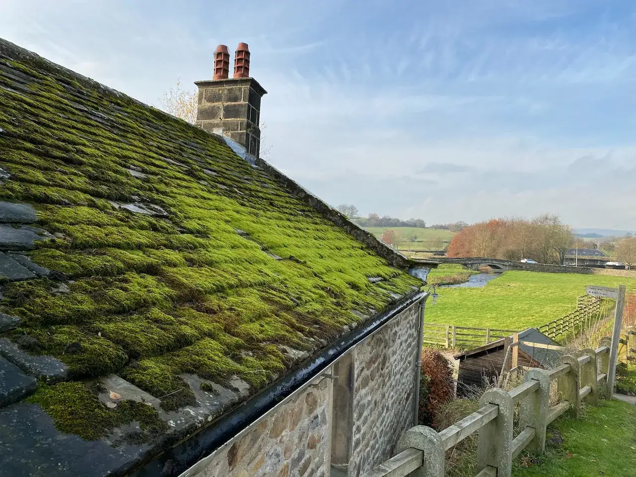 A stone cottage with a slate roof covered in vibrant green moss. A chimney with red pots stands tall against a blue sky. The countryside stretches out beyond.