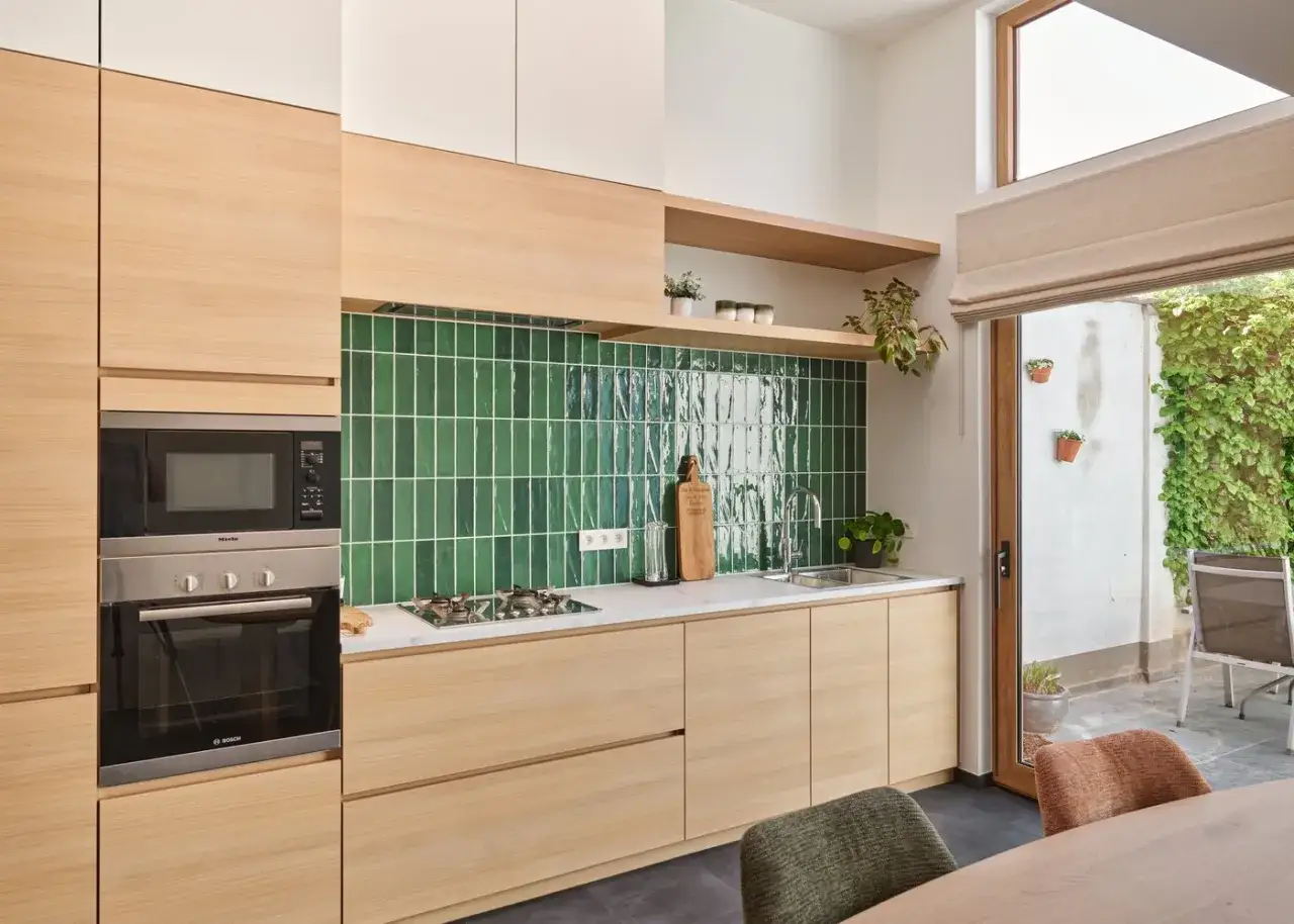 Cuisine moderne avec cr&eacute;dence verte brillante et meubles en bois clair. Un four et un micro-ondes int&eacute;gr&eacute;s. La **cuisine en parall&egrave;le** s'ouvre sur un patio verdoyant.