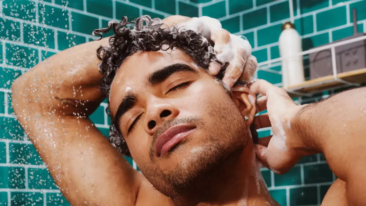 Hombre con ojos cerrados disfrutando de la higiene del cabello bajo la ducha, con espuma y gotas de agua.