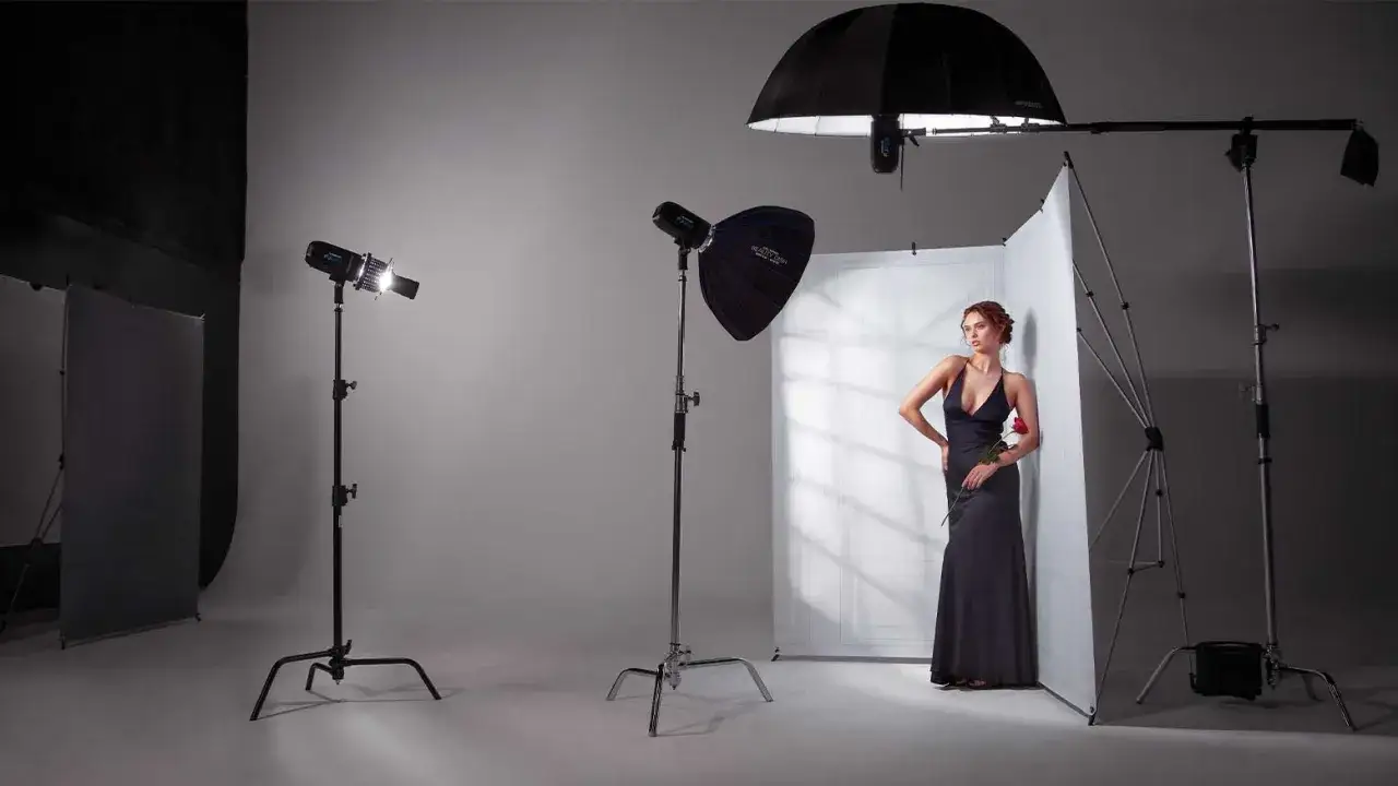 Modelka w ciemnej sukni pozuje przy białej ściance, otoczona przez profesjonalne modyfikatory światła: parasol, beauty dish i softbox.