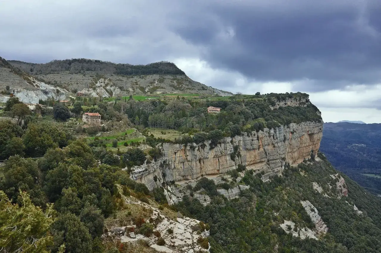 Paisaje montañoso con casas rurales y un acantilado imponente, como si fuera un coll sa cabra.