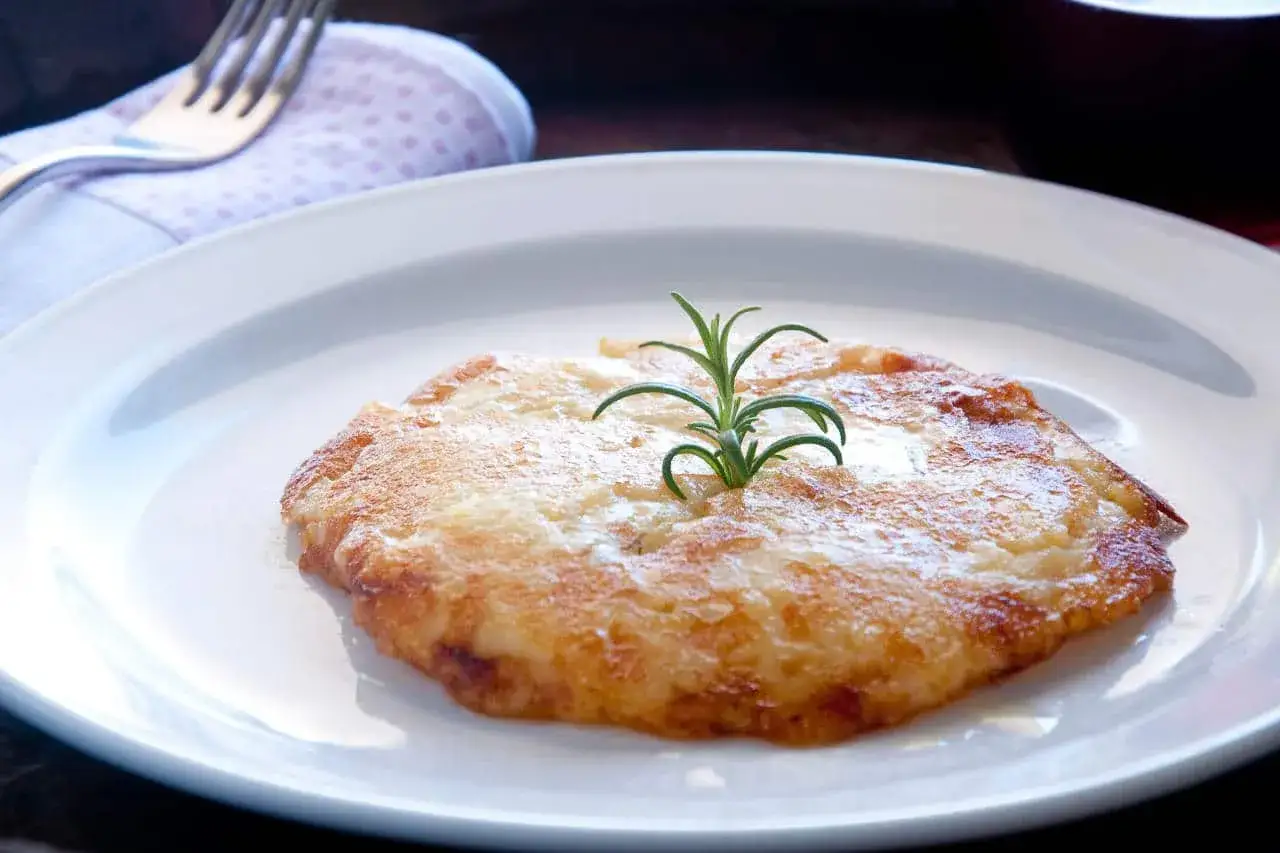 Piatto tipico udinese: frico dorato e croccante, guarnito con rosmarino fresco, servito su un piatto bianco.