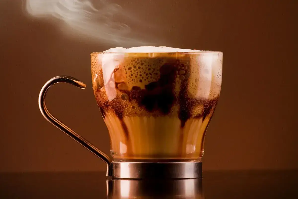 Un fumante caff&egrave; marocchino in una tazza di vetro con manico dorato, con schiuma e gocce di cioccolato.