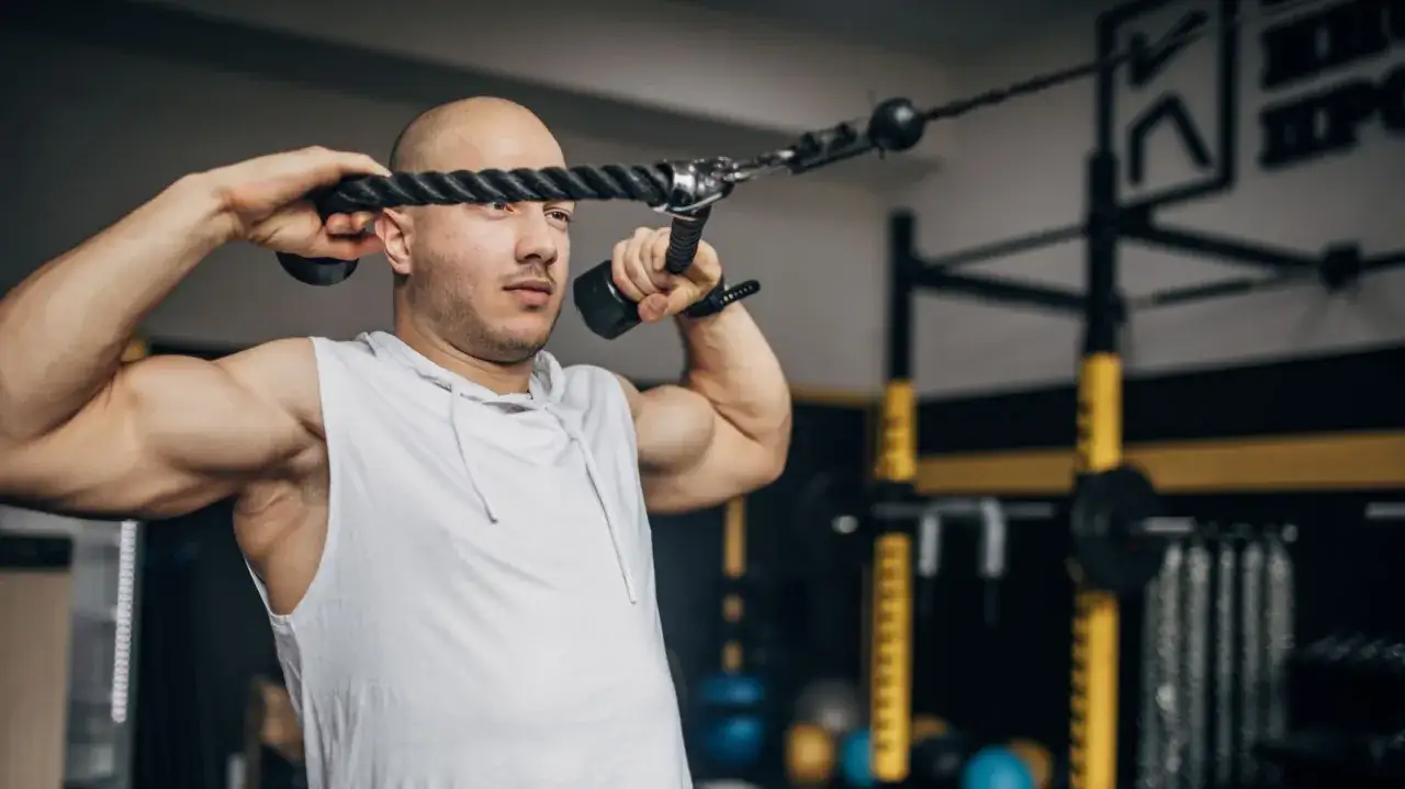 Man med rakad skalle utför face pulls med ett rep i ett gym.