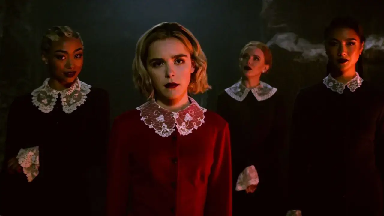 Chilling adventures of Sabrina obsada: cztery młode kobiety w czarnych strojach z koronkowymi kołnierzykami i jedna w czerwonym, stoją w mroku.
