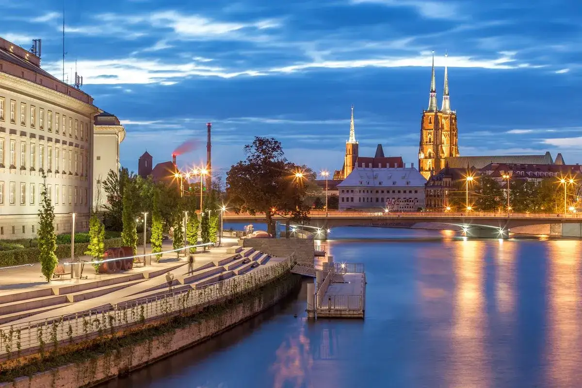 Wieczorny widok na Odrę i zabytkowe budowle Wrocławia, największego miasta zachodniej Polski. Oświetlone katedry i mosty odbijają się w wodzie.