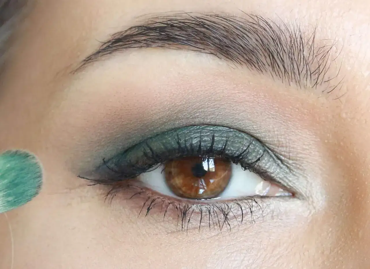 Occhi verdi marroni con un tocco di verde smeraldo sull'eyeliner, mentre un pennello applica il trucco.