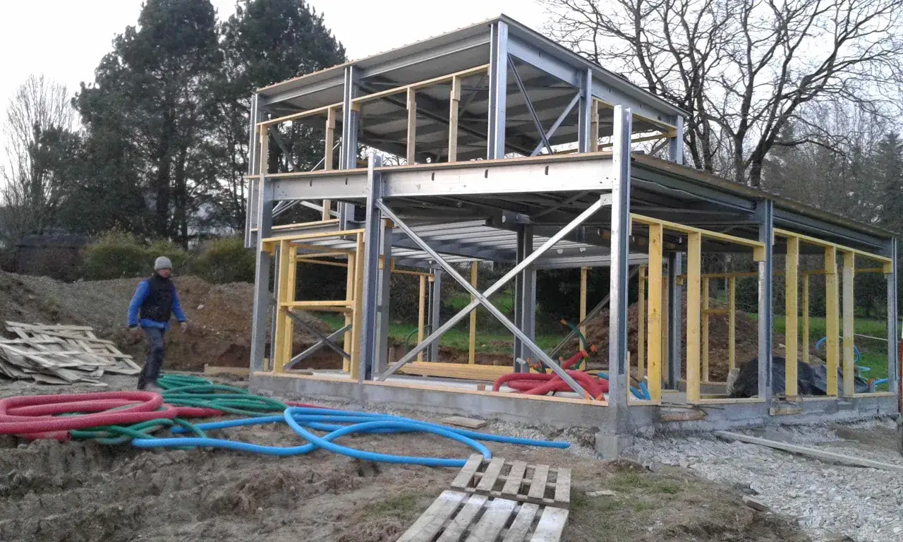Chantier d'une maison ossature m&eacute;tallique, structure en acier et bois, tuyaux color&eacute;s au sol.