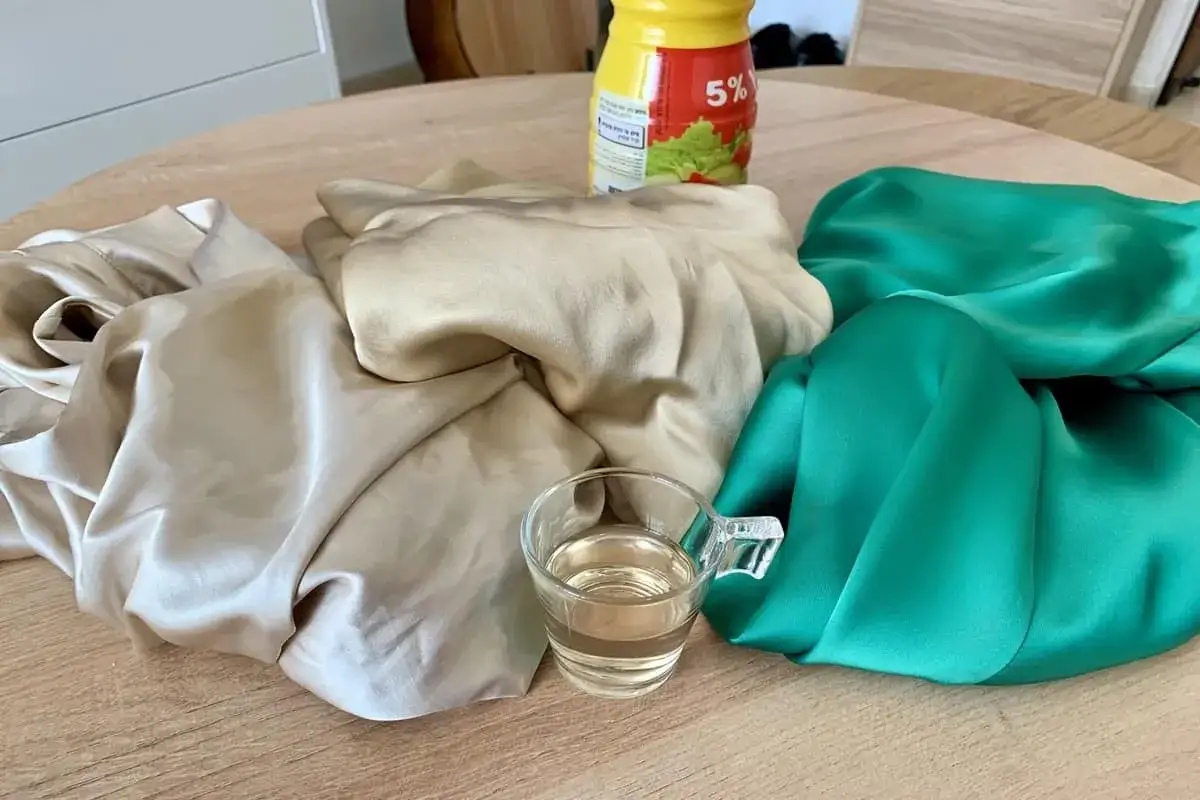 Tela de seda color crema y verde esmeralda junto a una taza con líquido claro, listo para lavar seda con vinagre.