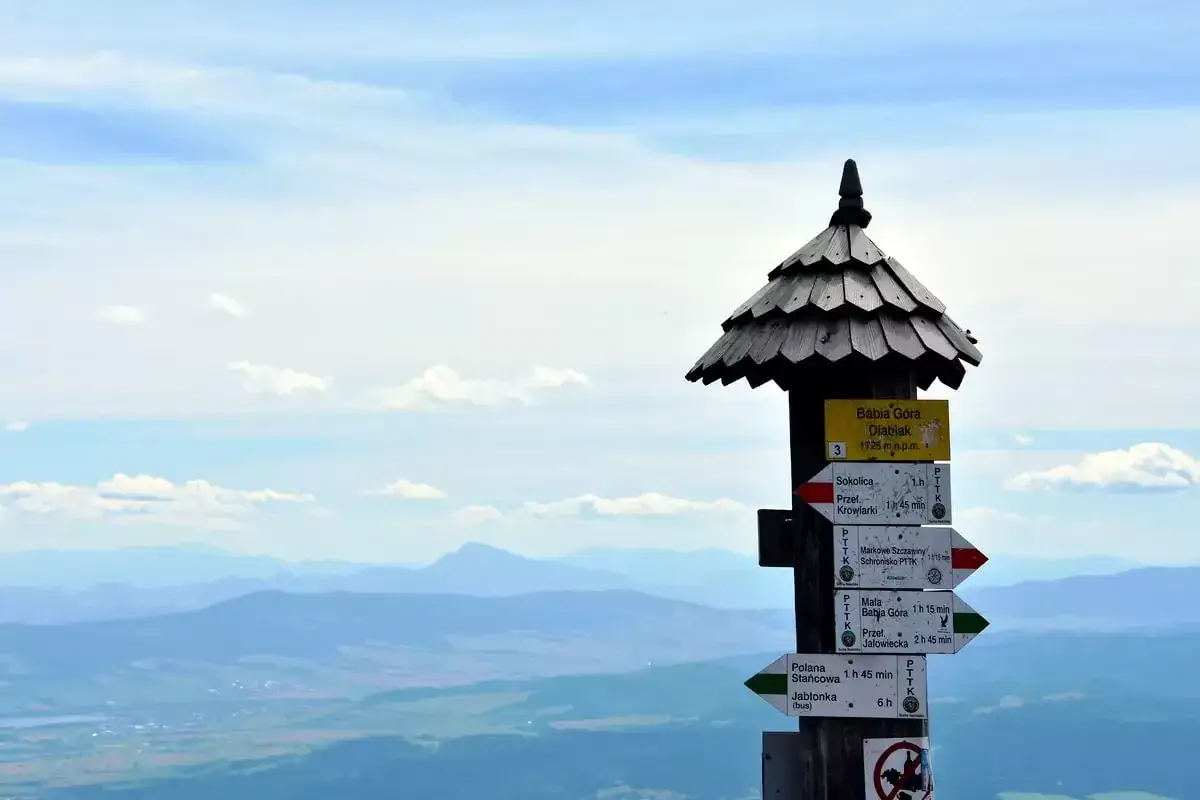 Słup z drogowskazami na szczycie, informujący ile ma Babia Góra Diablak (1725 m n.p.m.). W tle rozległy, górzysty krajobraz.