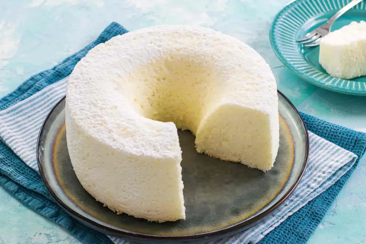 Una soffice angel cake altissima, spolverata di zucchero a velo, su un piatto scuro. Un pezzo è già stato servito.