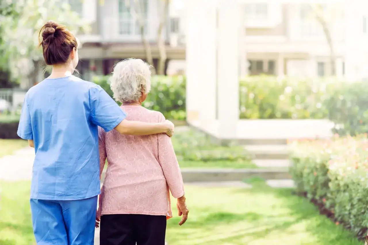Un'infermiera con una mano sulla spalla di un'anziana, passeggiano in un giardino. Un gesto di cura e supporto per chi vive con una persona ansiosa.