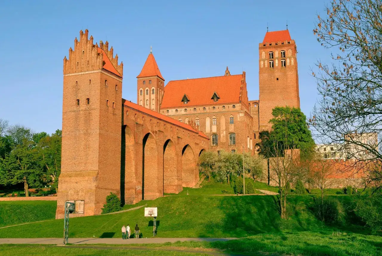 Potężny zamek w Kwidzynie z cegły, z charakterystycznymi wieżami i arkadowym gankiem, góruje nad zielonym wzgórzem.