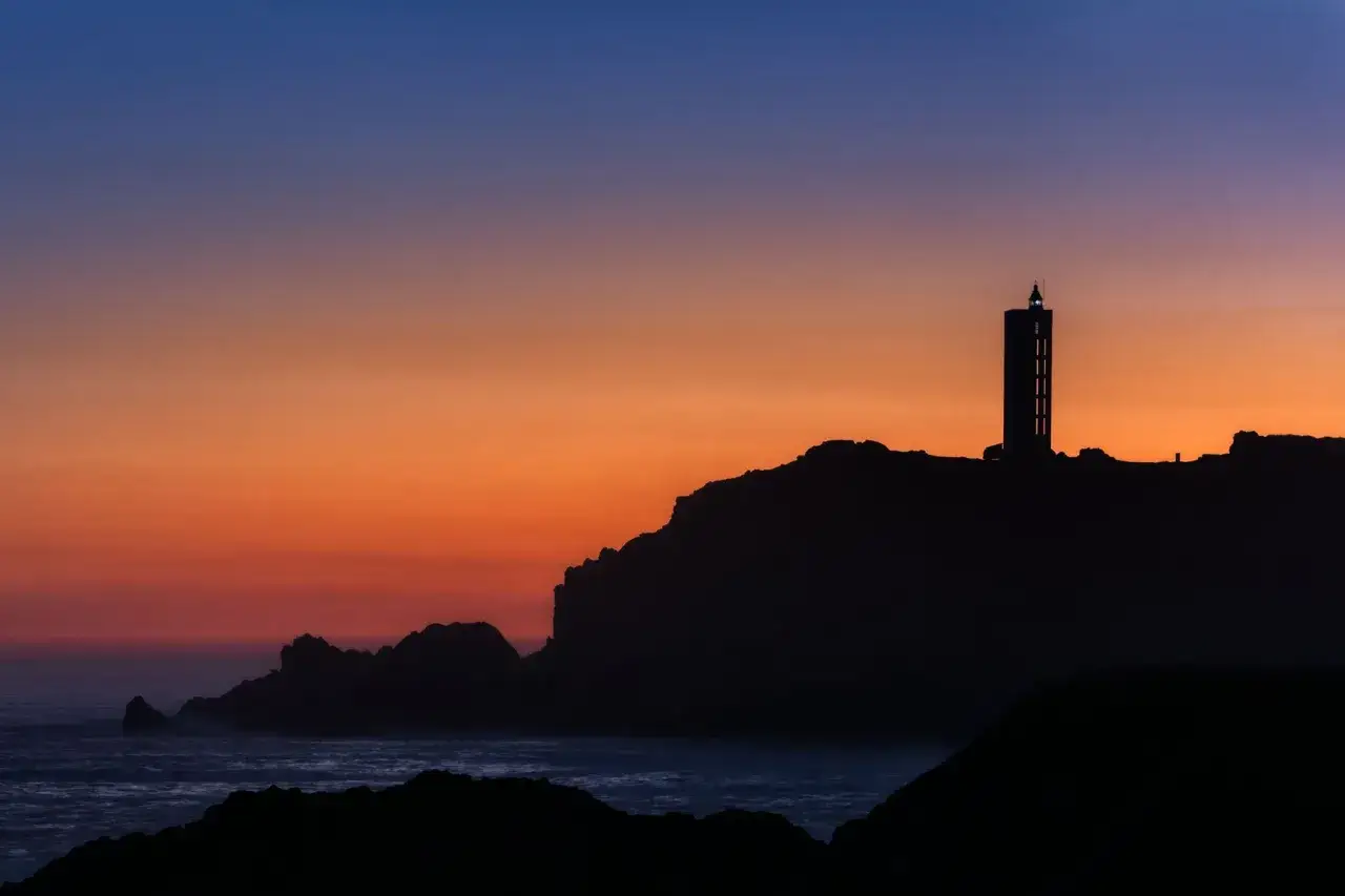 Silueta del faro de Punta Frouxeira contra un cielo anaranjado y azul al atardecer.