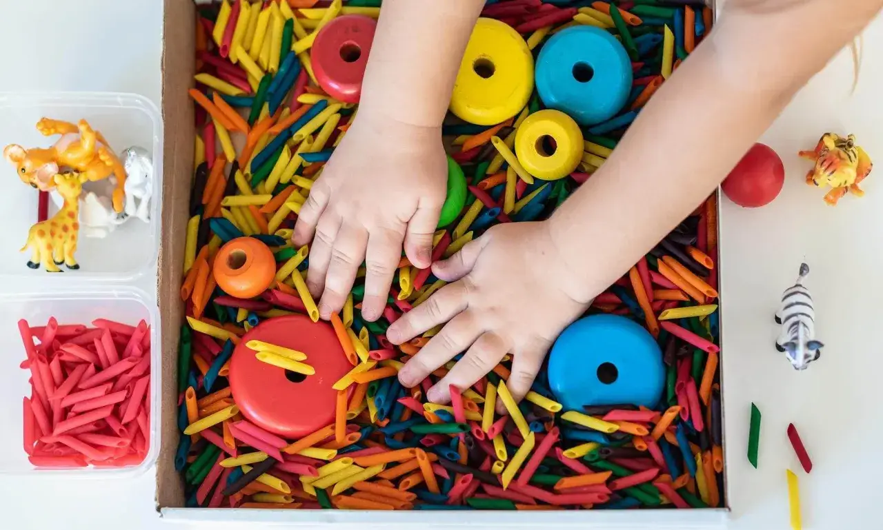 Manos de niño explorando un material sensorial de pasta de colores con aros de madera y figuras de animales.