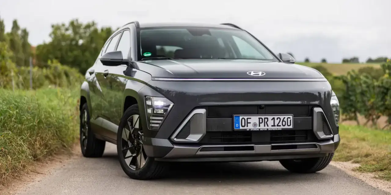 Der dunkle Hyundai Kona 2026 mit markanter Lichtleiste und LED-Scheinwerfern steht auf einer Landstra&szlig;e, umgeben von gr&uuml;ner Natur.