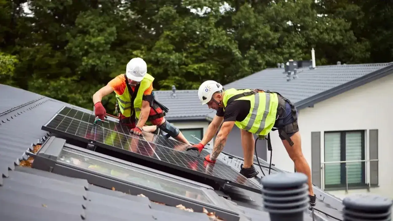 Zwei Handwerker mit Schutzhelmen und Warnwesten montieren Solarmodule auf einem Dach. Regelmäßige Wartung der Photovoltaikanlage ist Pflicht für optimale Leistung.