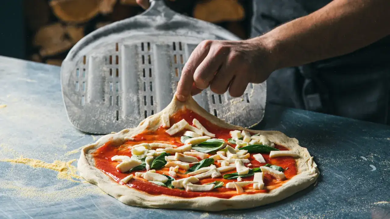 En hand lägger på ost på en pizza med tomatsås och basilika. En pizzaspade ligger bakom, redo för pizzaugnen. Perfekt för ditt pizza i pizzaugn recept.