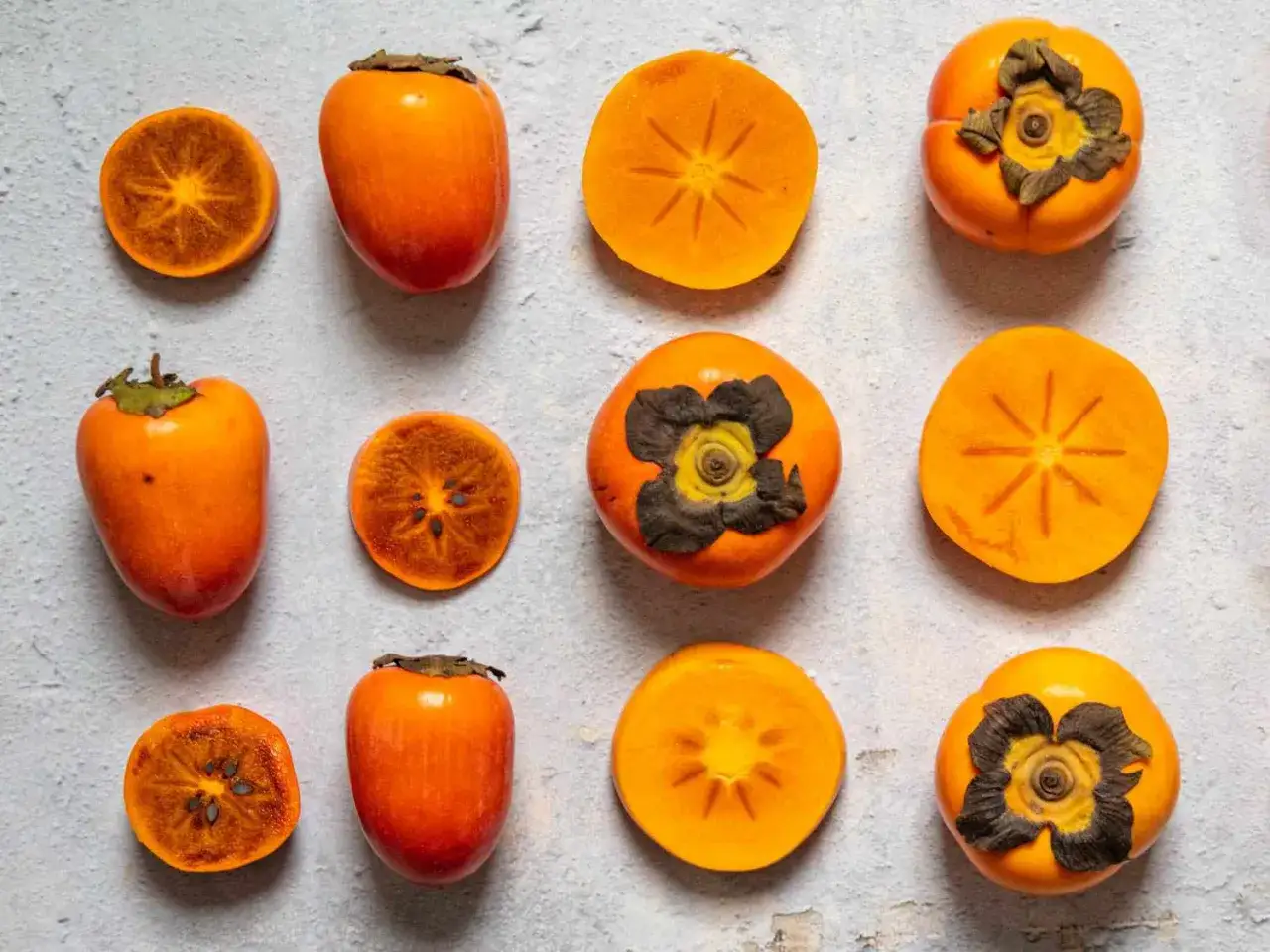 Mehrere ganze und halbierte Kaki-Fr&uuml;chte (kaki fruit in english) liegen auf einer hellgrauen, texturierten Oberfl&auml;che.