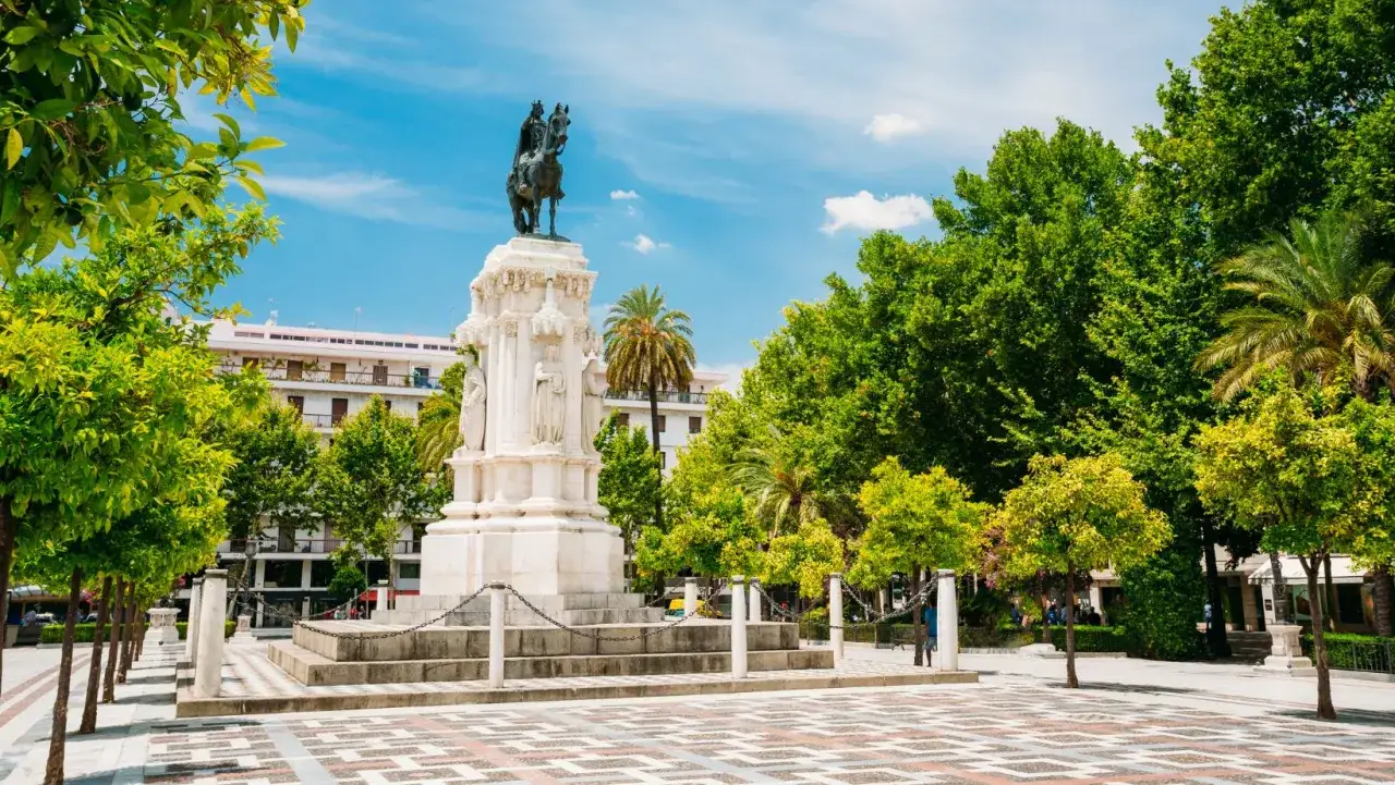 Majestuoso monumento ecuestre, una de las **estatuas en Sevilla**, rodeado de árboles frondosos y un cielo azul.