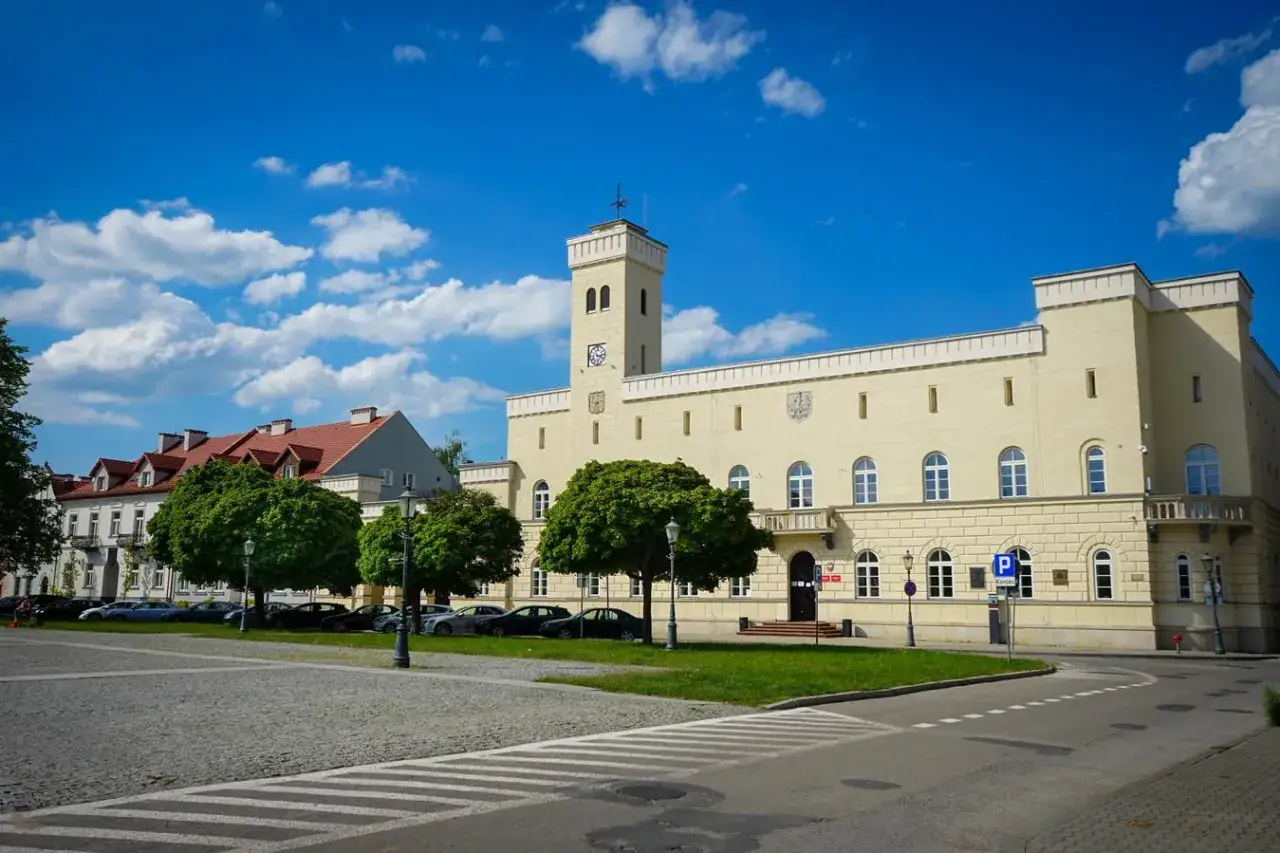 Jasny budynek ratusza z wieżą i zegarem, otoczony zielenią i brukowanym placem. Jedna z radomskich atrakcji.