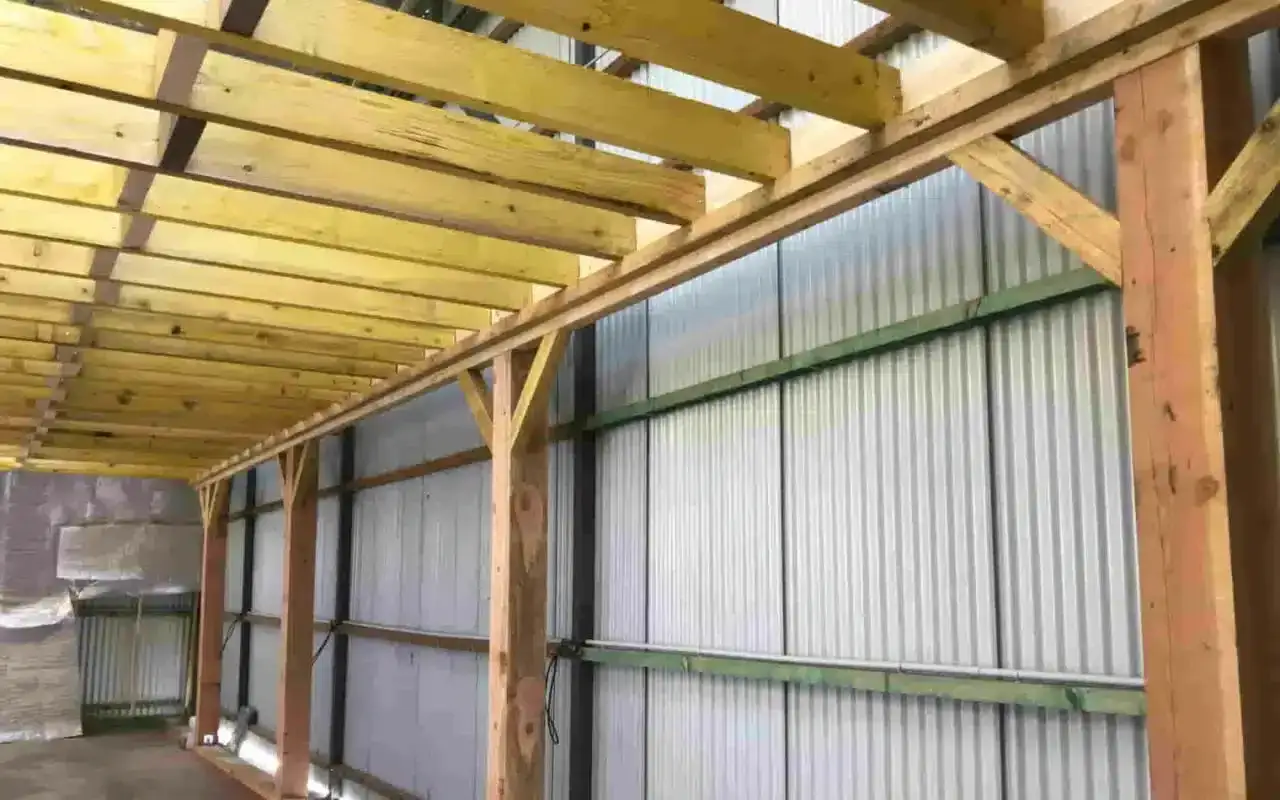 Structure en bois avec des madriers ou bastaings supportant un toit. Le mur est en t&ocirc;le ondul&eacute;e.