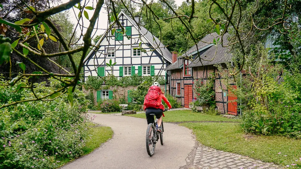 Radfahrerin mit rotem Rucksack fährt auf einem Weg an Fachwerkhäusern vorbei. Ein idyllisches Bild für eine Fahrradtour in NRW.