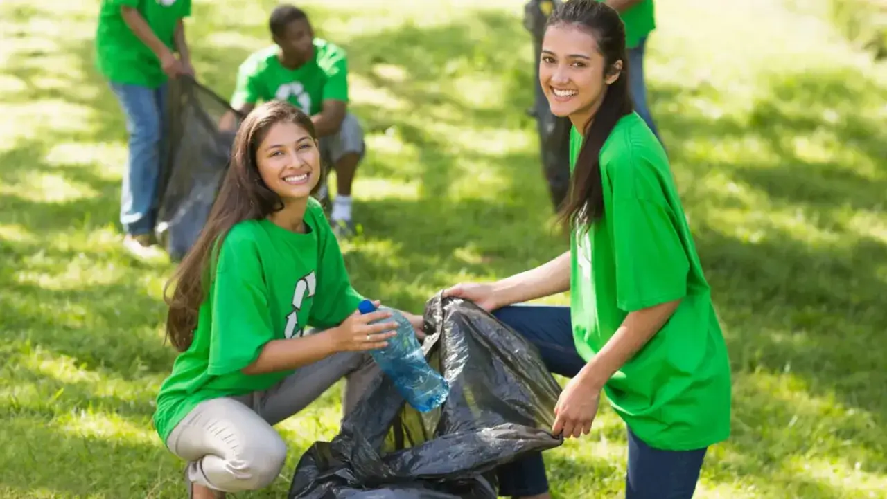 Two smiling young women in green shirts demonstrate volunteer skills by cleaning up a park, collecting trash in a bag.