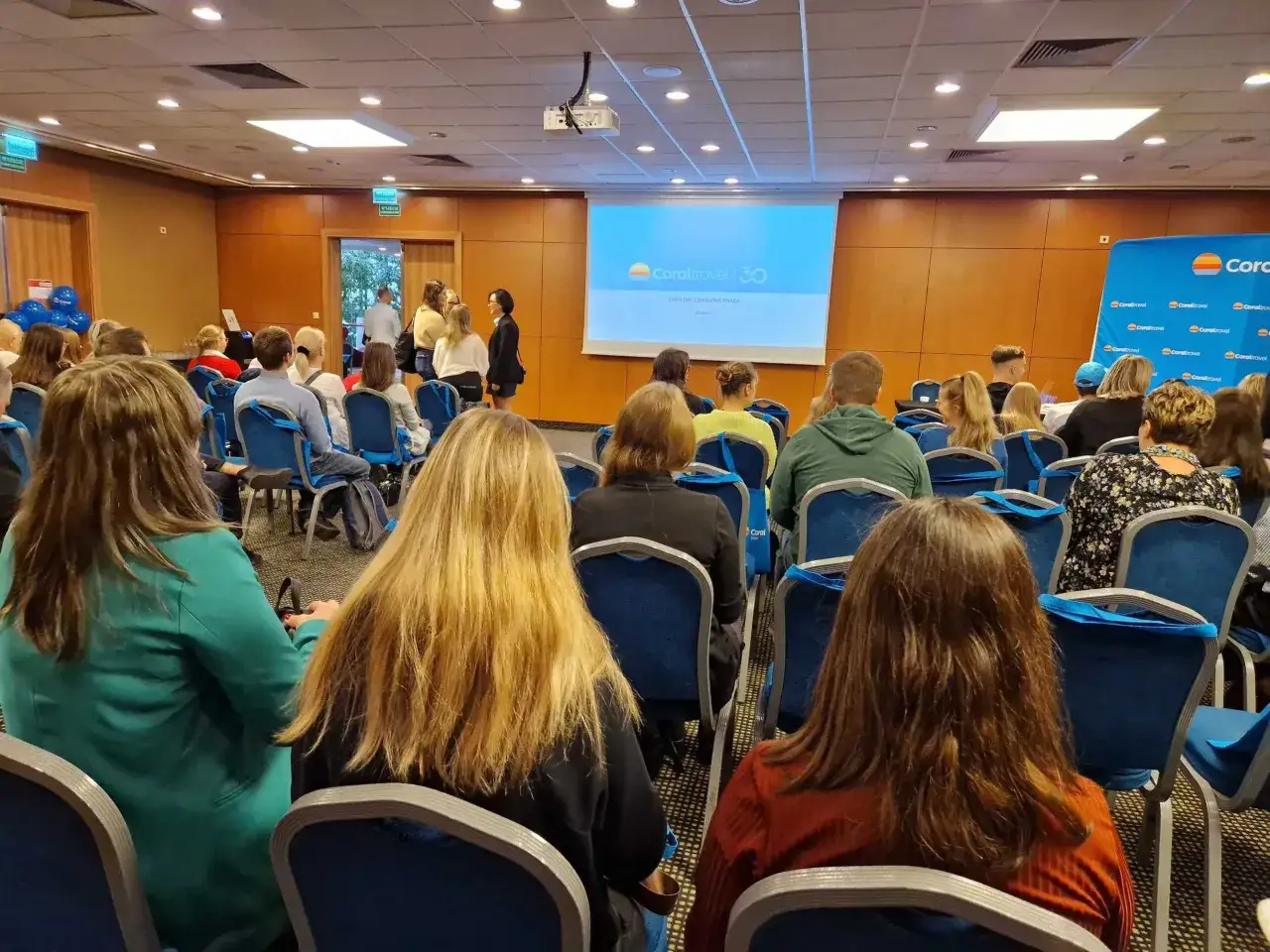 Ludzie siedzą na sali konferencyjnej, słuchając prezentacji. Na ekranie logo Coral Travel, a obok stoi rezydent.