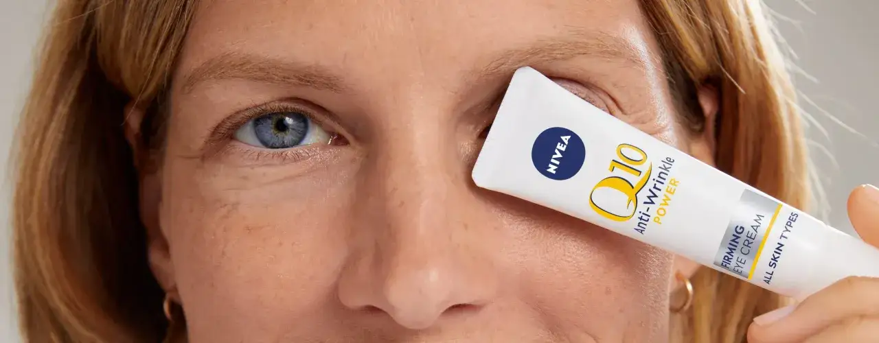 Un tube de crème contour des yeux Nivea Q10 Power est tenu devant l'œil d'une femme, camouflant les rides pattes d'oie.