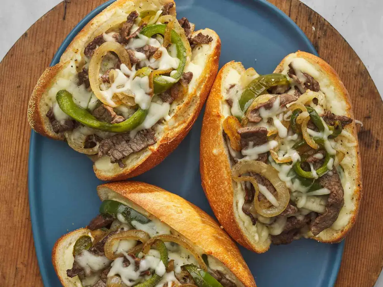Tre saftiga philly steak sandwiches med smält ost, paprika och lök på en blå tallrik.