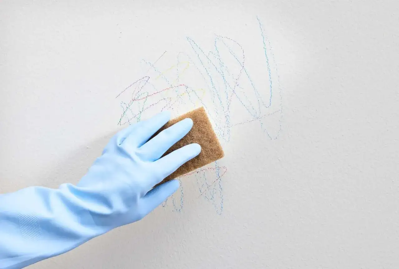 Mano con guante azul limpiando ceras de colores de una pared blanca con una esponja.