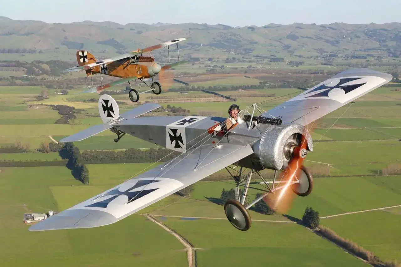 Zwei historische Fokker Eindecker fliegen &uuml;ber gr&uuml;ne Felder. Ein Pilot im Vordergrund winkt.