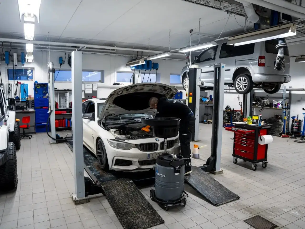 Mekaniker arbetar med en vit BMW p&aring; en bilverkstad. En annan bil h&auml;nger i en lyft. H&auml;r ser du vad som ing&aring;r i bilservice.
