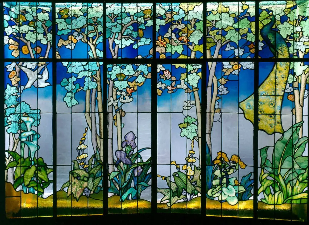 Vitrail de Jacques Gruber : un paon majestueux et un colvert s'épanouissent dans un jardin luxuriant, sous un ciel bleu éclatant.