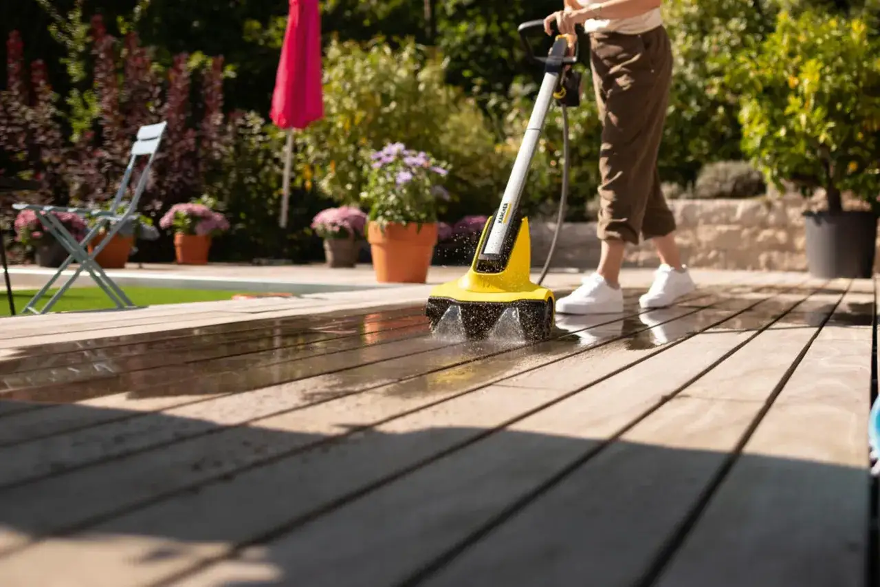 Eine Person reinigt mit einem K&auml;rcher die Bankirai-Terrasse. Wasser spritzt, w&auml;hrend die B&uuml;rste &uuml;ber das Holz gleitet.