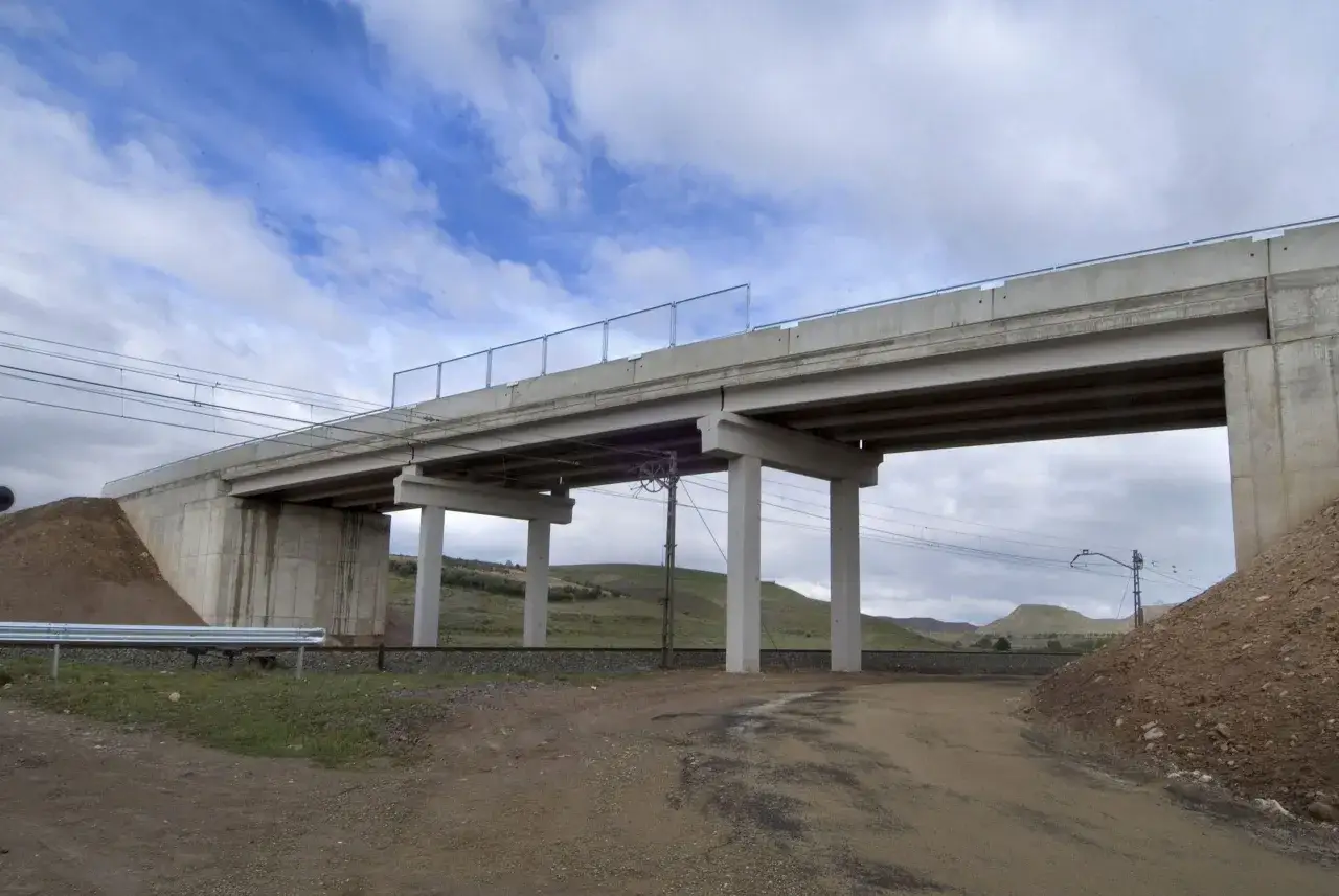 Un puente de hormigón se alza sobre vías de tren. Los pilares de este estribo soportan la estructura, mientras el cielo nublado se refleja en el paisaje.