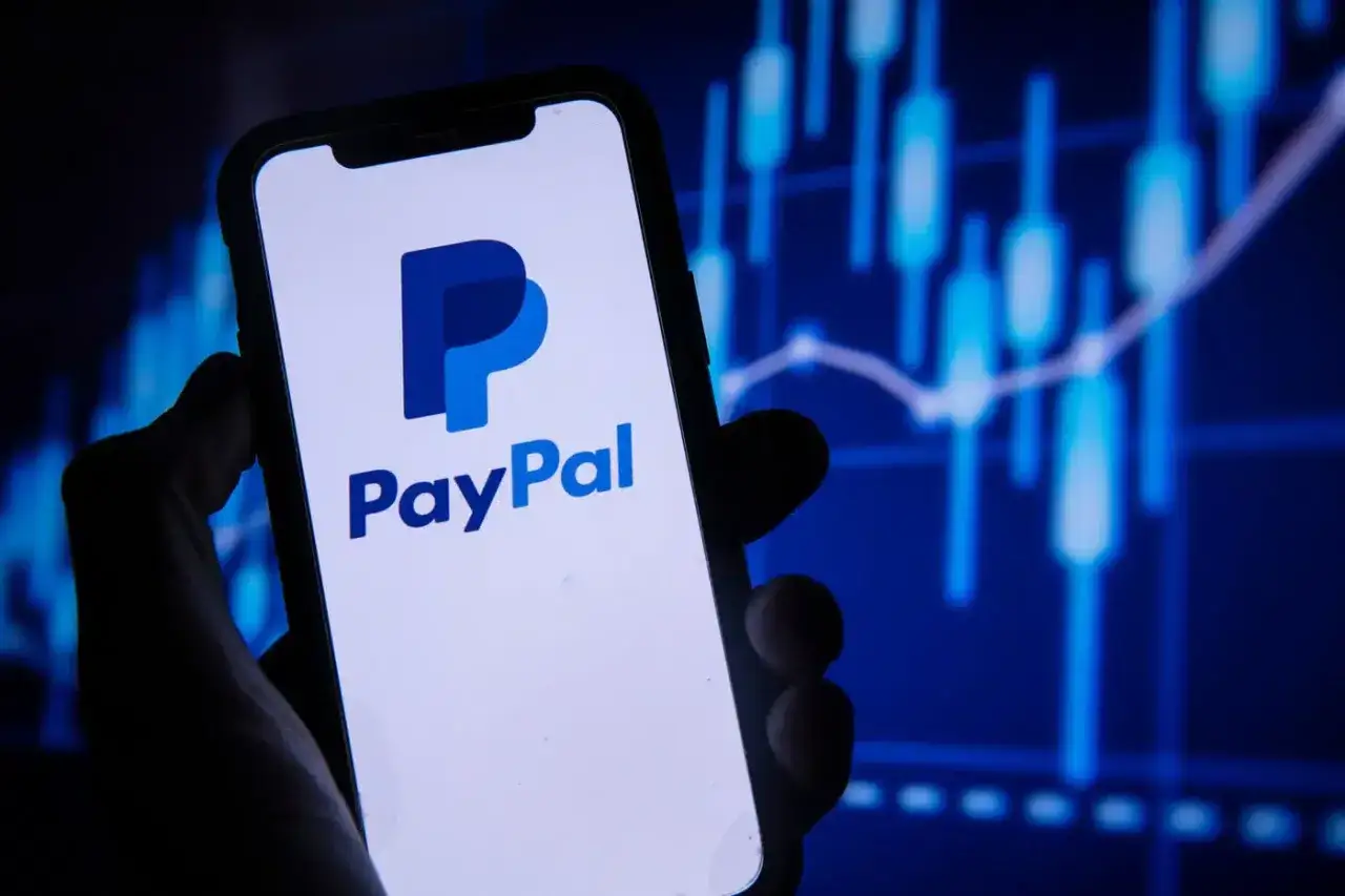 Dłoń trzyma smartfon z logo PayPal. Tło to wykres finansowy, sugerujący jak usunąć PayPal.