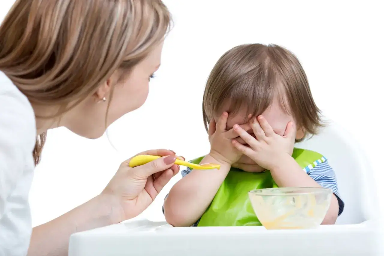 Mama próbuje nakarmić niejadka, ale maluch zakrywa buzię rączkami.