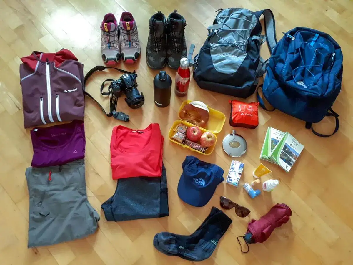 Packliste Wanderurlaub: Wanderschuhe, Rucksack, Kleidung, Kamera, Verpflegung, Trinkflasche, Sonnenbrille und Regenschirm für das nächste Abenteuer.