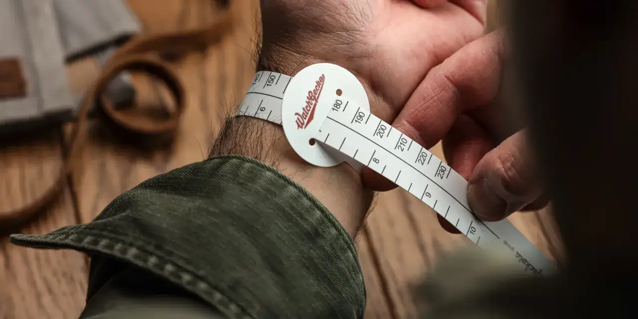 Mężczyzna mierzy nadgarstek taśmą, aby określić rozmiar zegarka. Widoczne są liczby od 140 do 230.