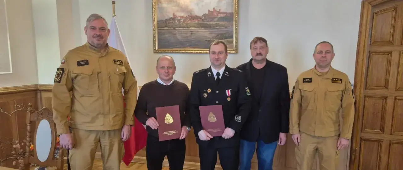 Pięciu mężczyzn pozuje do zdjęcia. Dwóch w mundurach strażackich, jeden w garniturze, a dwóch w beżowych kurtkach. Zastępca naczelnika OSP trzyma dokument.