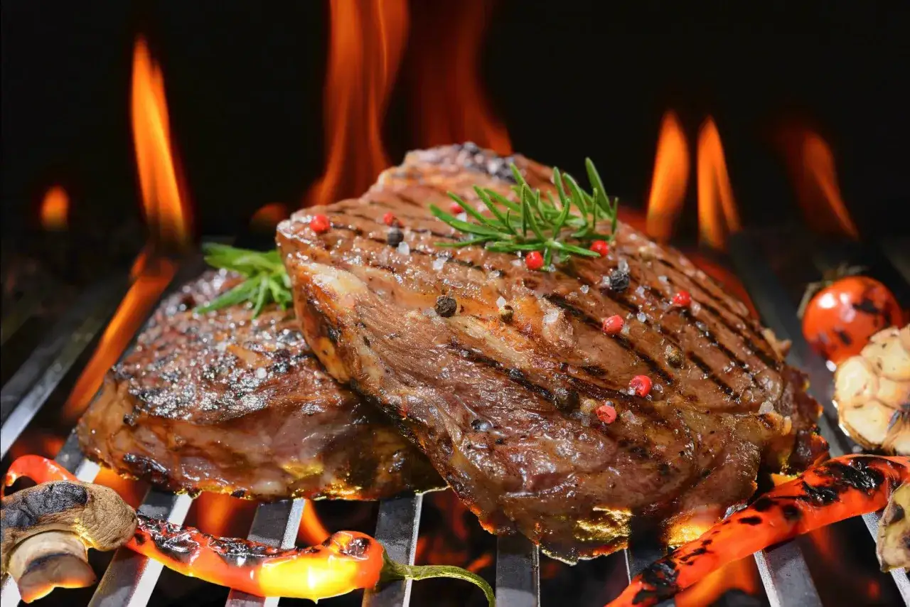 Dos jugosos filetes de carne asada, sazonados con sal y pimienta, se cocinan a la parrilla con llamas naranjas, acompañados de pimientos y tomates asados.