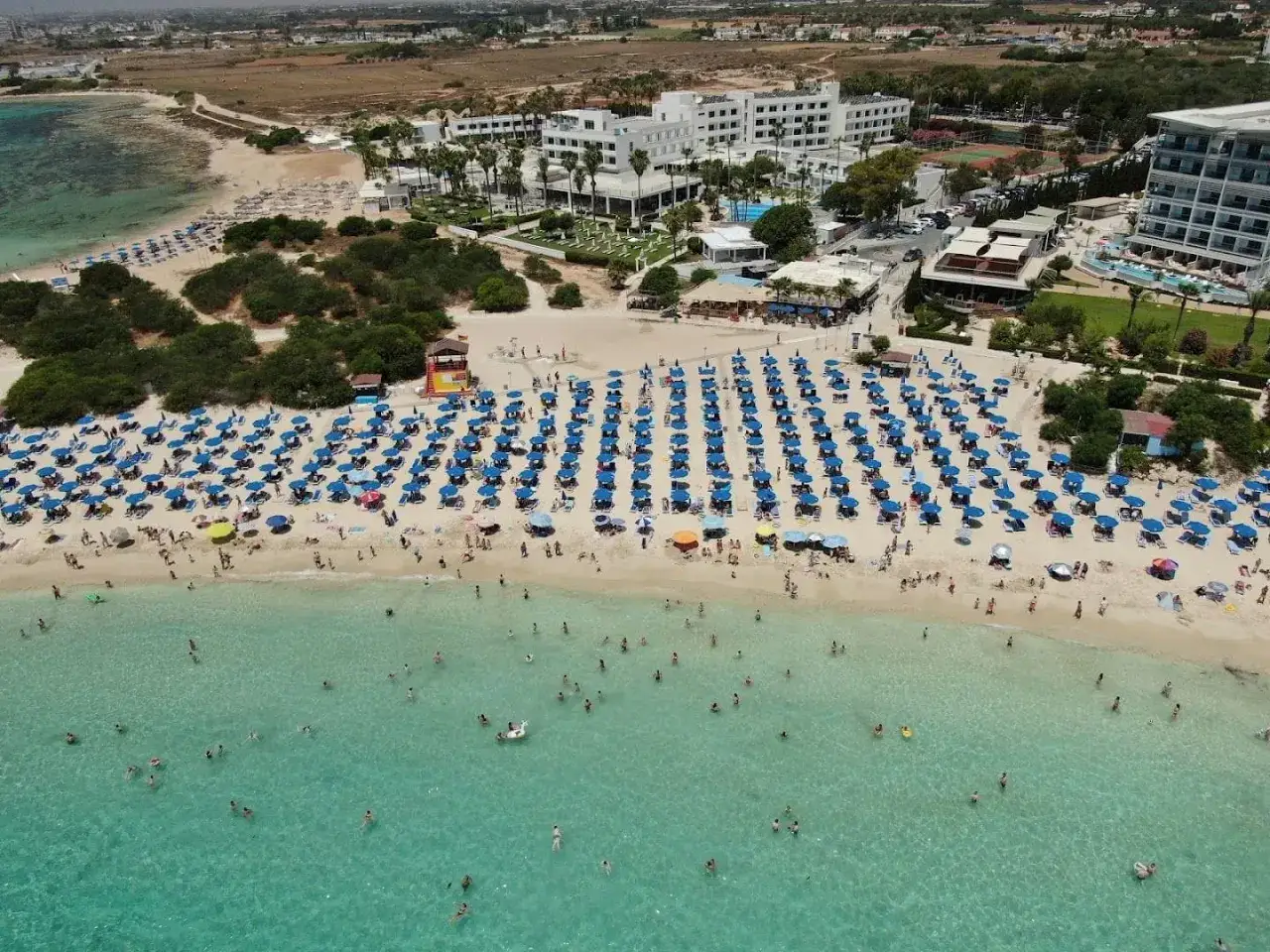 Tłum ludzi cieszy się słońcem na plaży Makronissos, z rzędami niebieskich parasoli i turkusową wodą.