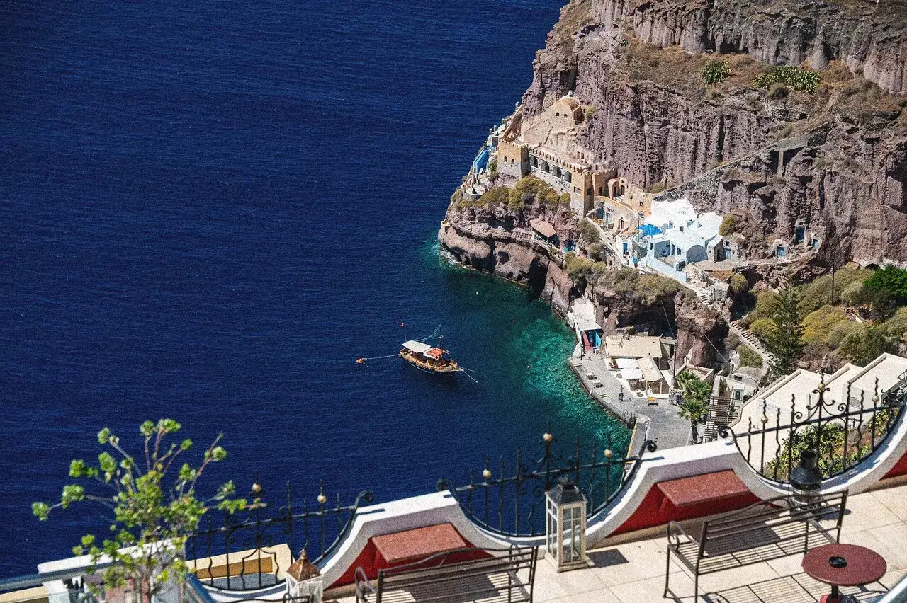 En båt guppar i det turkosa vattnet vid en klippig kust med vita hus. En perfekt plats att fira Santorini på.
