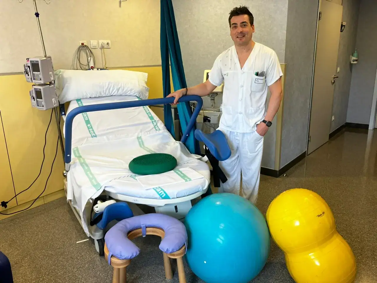 Médico sonriente junto a cama de parto y balones para partos naturales.