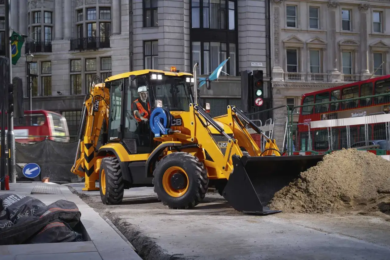 Ż&oacute;łta koparko-ładowarka JCB 3CX Compact pracuje na ulicy, ładując piasek. W tle widać zabytkową architekturę i czerwony autobus.