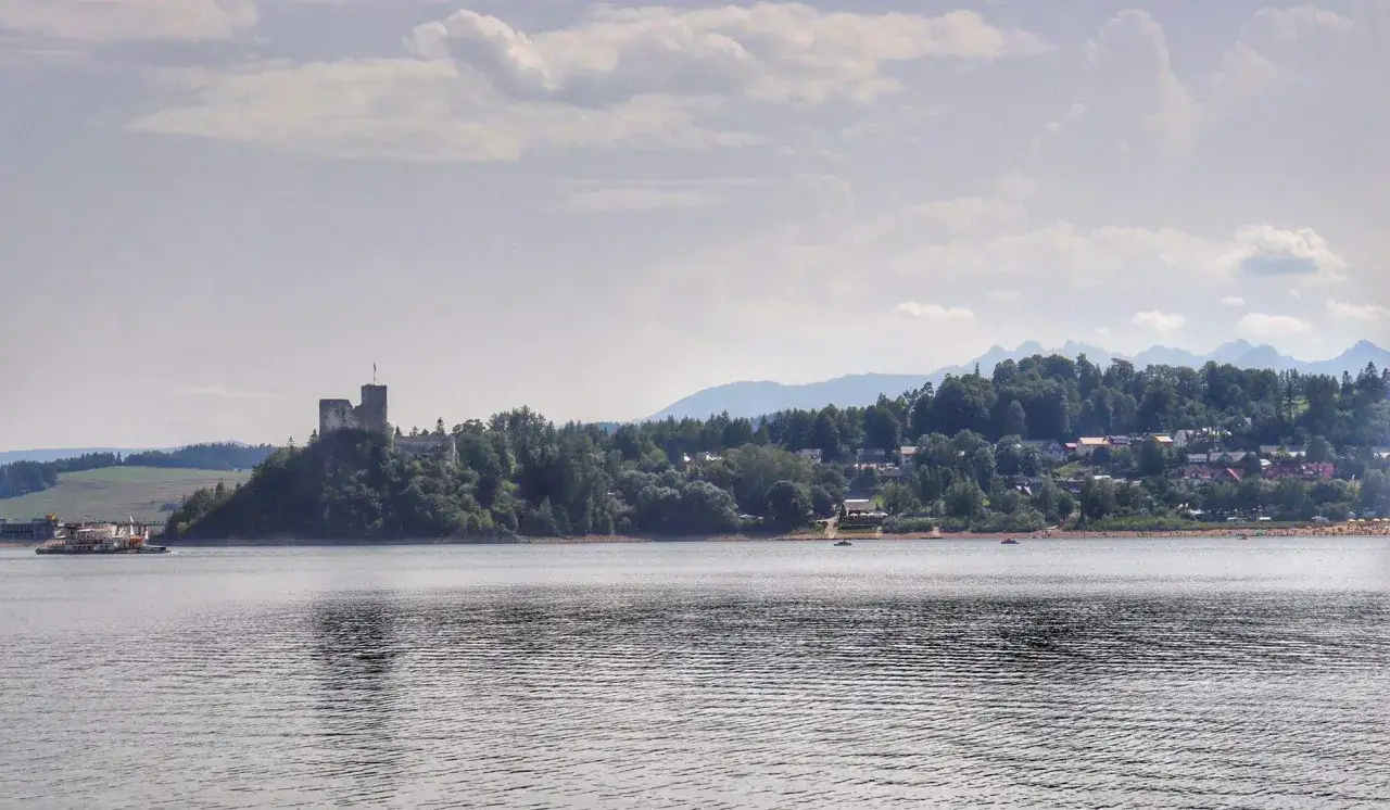 Zamek na wzgórzu nad spokojnym jeziorem Czorsztyńskim, z łodzią na wodzie i górami w tle.