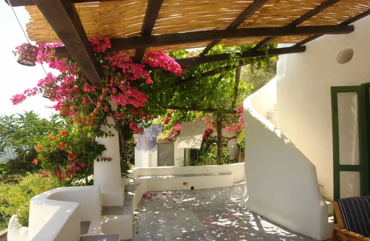Veranda aperta con bouganville rosa, rampicanti verdi e lanterne in bambù. Un esempio di come abbellire una veranda con piante e luci naturali.