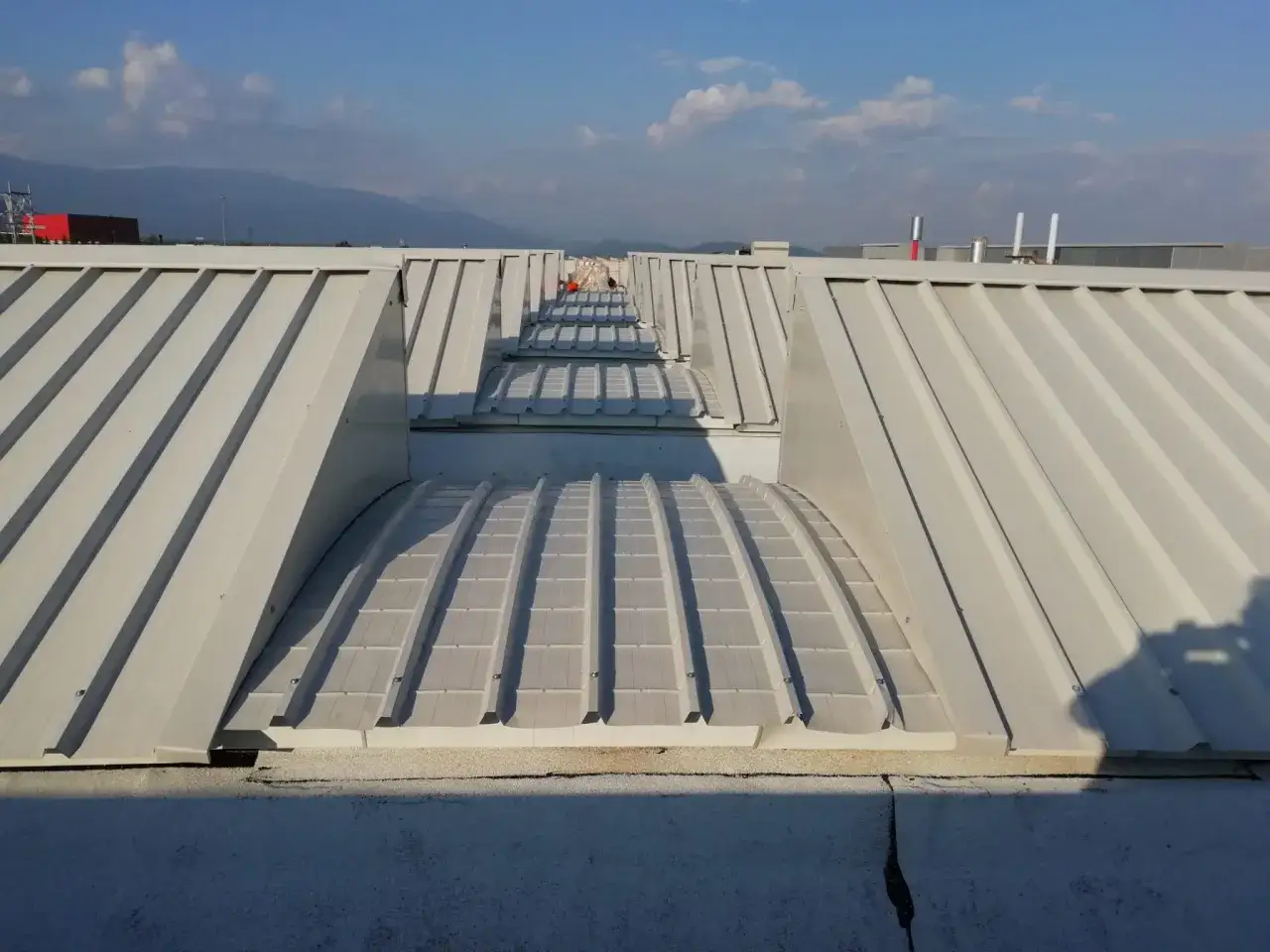 Coperture industriali bianche con lucernari a botte, sotto un cielo azzurro con nuvole bianche.